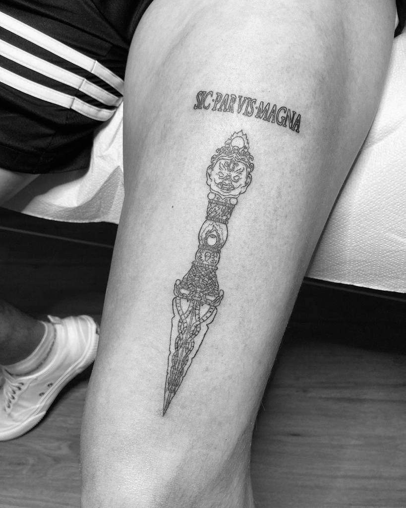 Black and white tattoo of a dagger with a face on a thigh, accompanied by the text "Sic Parvis Magna"