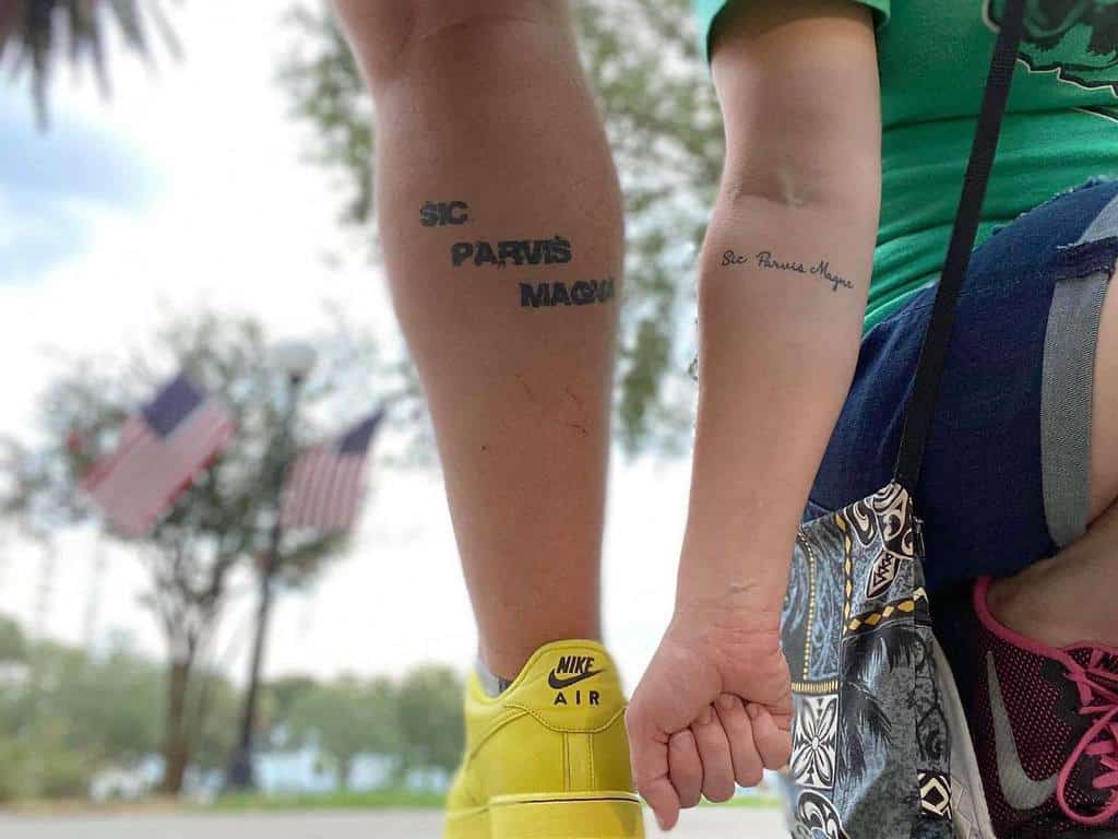 Two people showing matching tattoos on arm and leg with the phrase "Sic Parvis Magna"