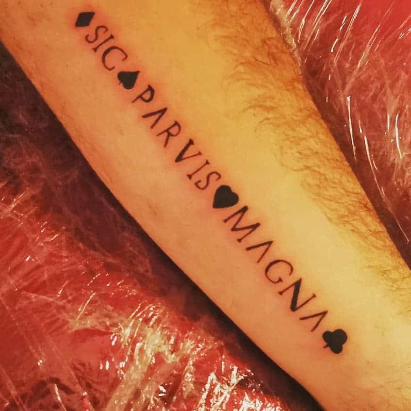 A tattoo on an arm reads "Sic Parvis Magna" with hearts and spade symbols between the words