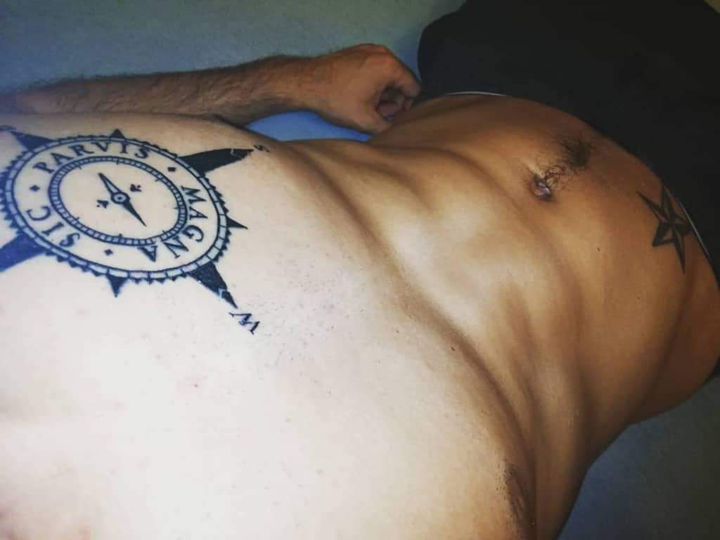 Abdomen tattoo featuring a bold compass design with 'Sic Parvis Magna' text in a prominent black ink style