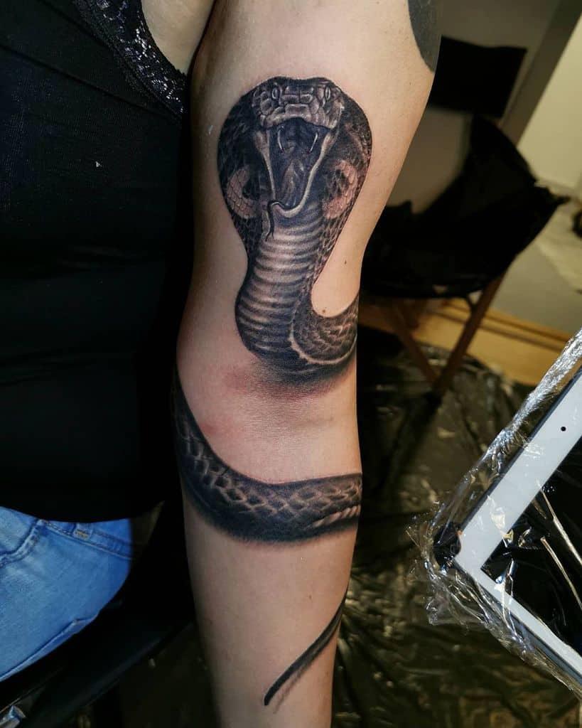 Tattoo of a coiled cobra on a person's arm, with mouth open and hood expanded