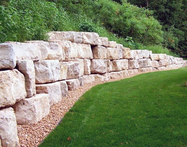 Large natural stone retaining wall with gravel base and a sloped green lawn.