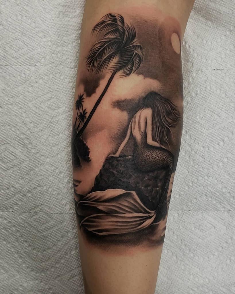 Black and grey tattoo of a mermaid sitting on a rock under a moonlit sky, with palm trees and ocean waves creating a serene scene.