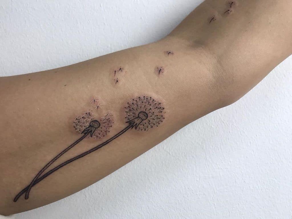 This tattoo features two dandelions, with the delicate seeds appearing to drift away, showcasing the beauty of movement and fragility in nature. The design captures a soft, whimsical touch.