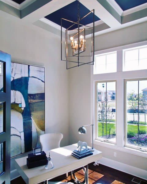 Bright home office with white desk, abstract art, large windows, and modern chandelier.