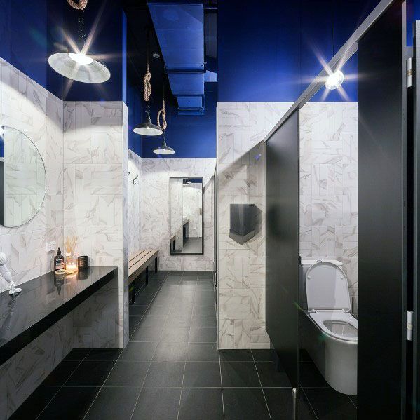 Modern restroom with black partitions, white marble walls, blue ceiling, and industrial lighting.