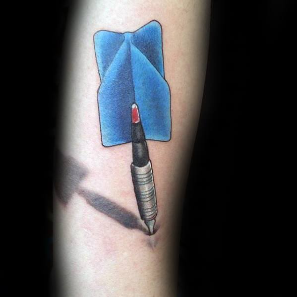 Color forearm tattoo of a dart piercing the skin with a bold blue flight and realistic shadow, symbolizing precision and passion.