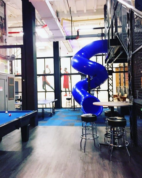 Modern indoor space with a blue slide, pool table, high stools, and sports murals