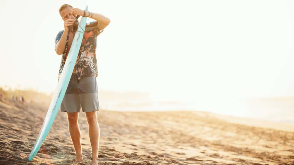 A man on the beach in board shorts attaching something to a surfboard