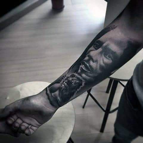 Bob Marley portrait tattoo with a lion below, inked in black and gray realism style on the inner forearm.