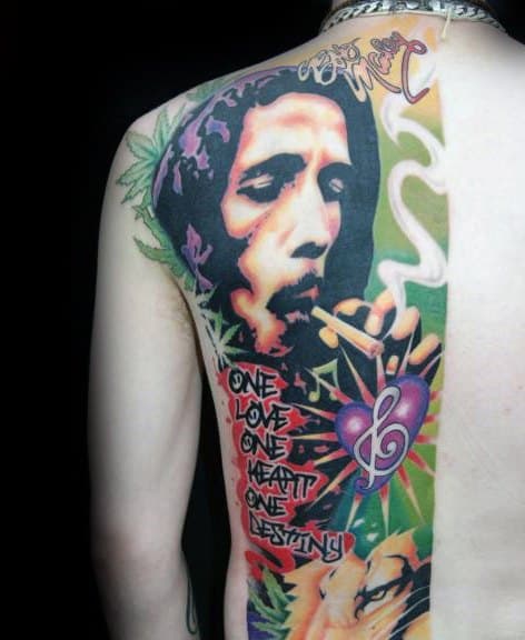 Vibrant upper back tattoo of Bob Marley smoking, with colorful elements like a lion, cannabis leaves, and the quote “One Love One Heart One Destiny.”