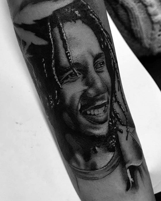 Black and gray tattoo of Bob Marley smiling with braided hair, inked on the forearm in a lifelike portrait style.