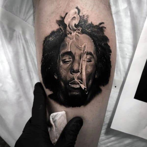 Realistic tattoo of Bob Marley with closed eyes and smoke rising from a joint, centered on the calf with soft shading and lifelike texture.