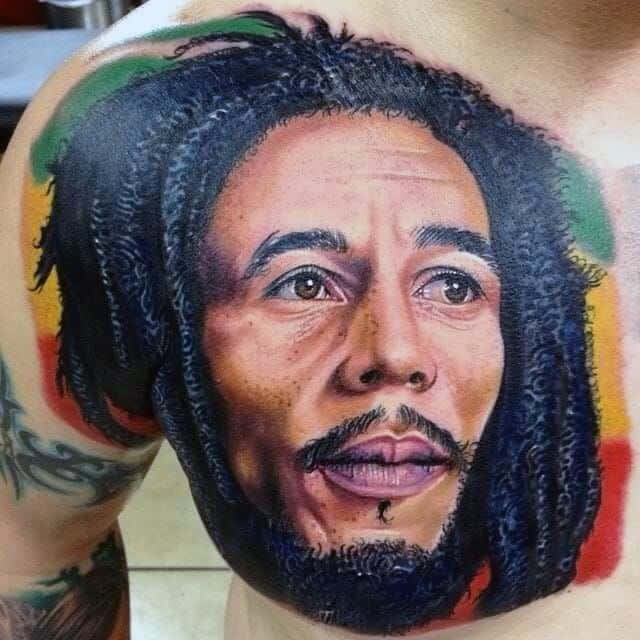 Vivid shoulder tattoo of Bob Marley in hyper-realistic color, with red, yellow, and green background accents symbolizing Rastafarian culture.
