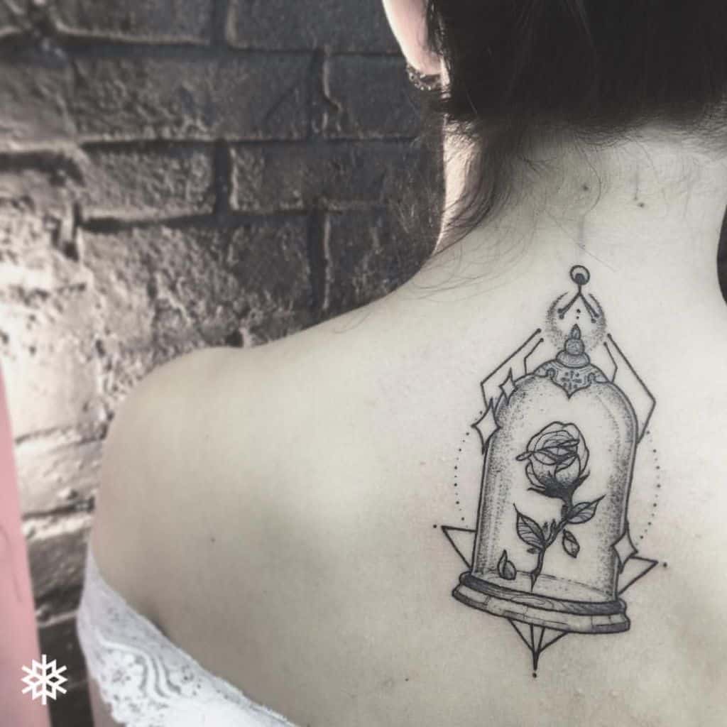 Elegant black ink tattoo of the enchanted rose inside a glass dome, placed on the upper back with fine-line details and geometric accents.