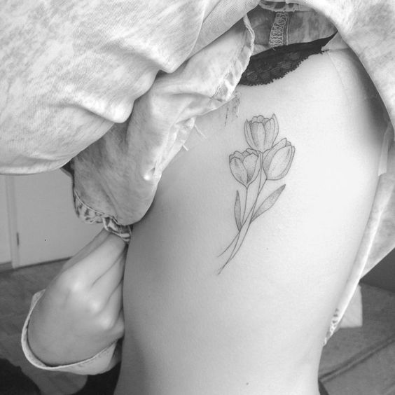 A delicate black and grey tulip tattoo on the ribcage, featuring three blooming flowers with fine shading and soft, elegant line work.