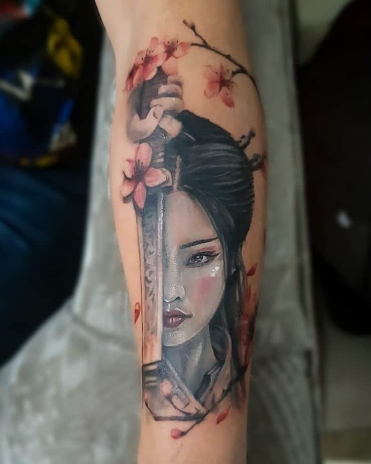 Realistic forearm tattoo of a Geisha holding a katana, with her face split by the blade, surrounded by cherry blossoms and soft shading.