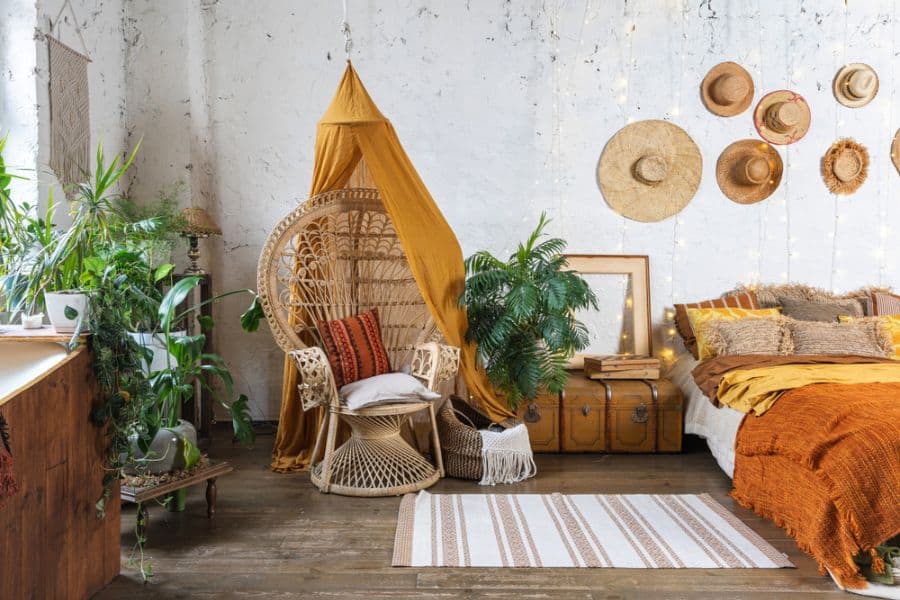 Boho bedroom with wicker chair, wall hats, plants, and cozy bedding in earthy tones.