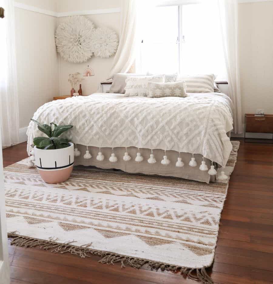 Cozy bedroom with white bedding, a tassel throw, potted plant, and decorative wall hangings on wooden floors