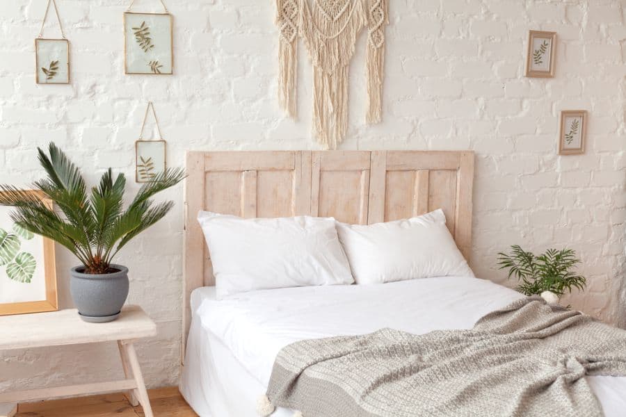 Minimalist bedroom with white bedding, wooden headboard, macrame, white potted plants, and framed leaf prints