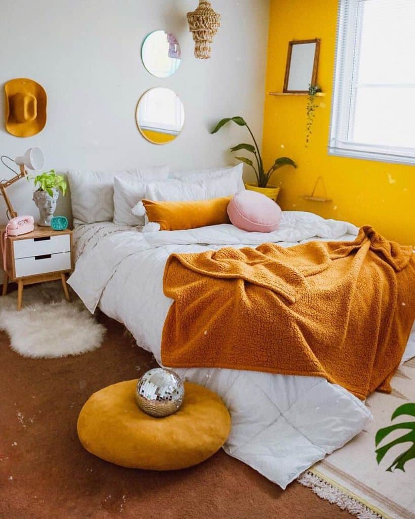 Cozy bedroom with a yellow accent wall, white bedding, orange throw, and round yellow mirrors