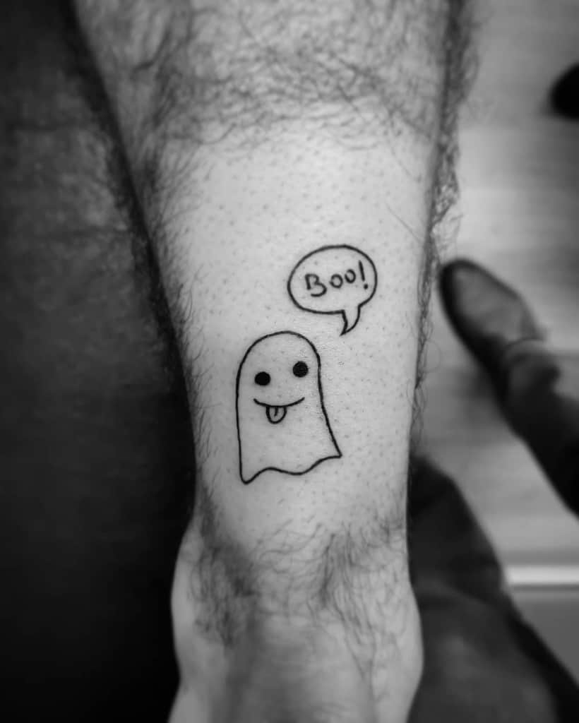 A simple black ink tattoo of a cute ghost sticking out its tongue, saying 'Boo!' in a speech bubble, designed in a playful and minimal style.