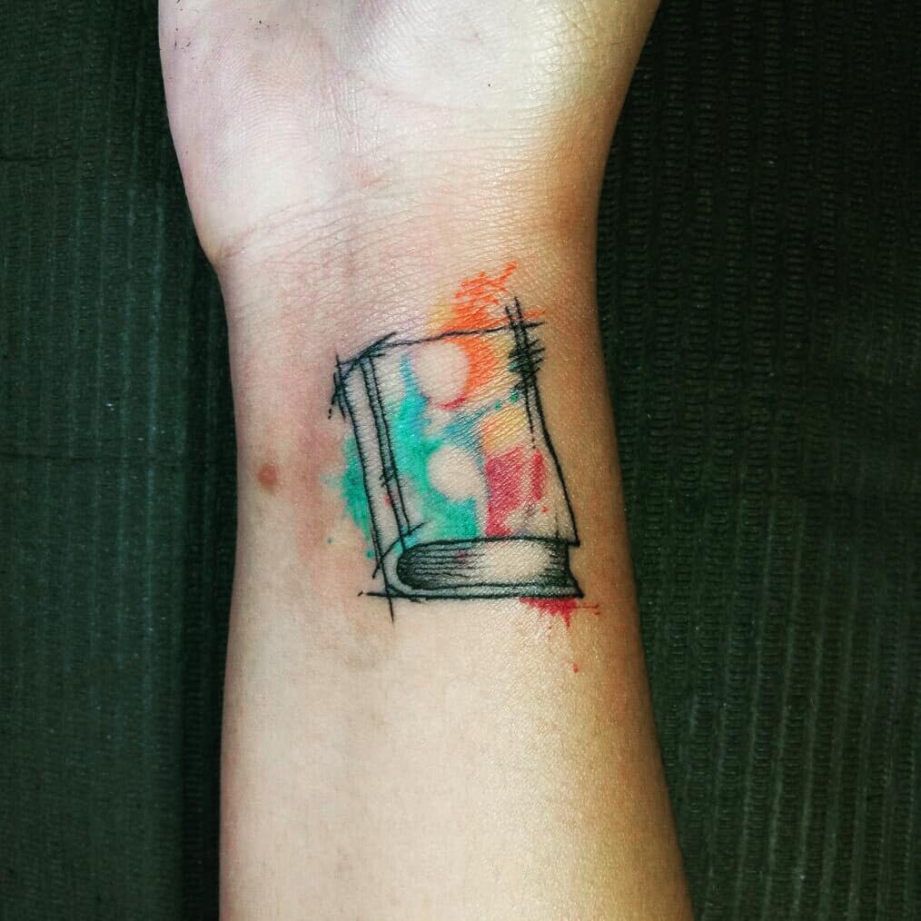This tattoo features a book with an abstract watercolor design, offering a creative and colorful interpretation. The lines and watercolor splashes add a dynamic and artistic touch to the classic concept of a book.