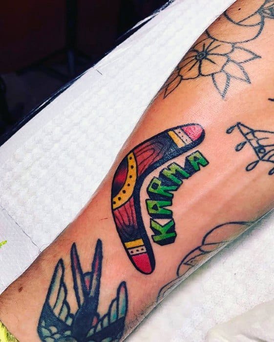 Colorful boomerang tattoo with tribal patterns and the word KARMA in bold green letters, placed on the forearm.