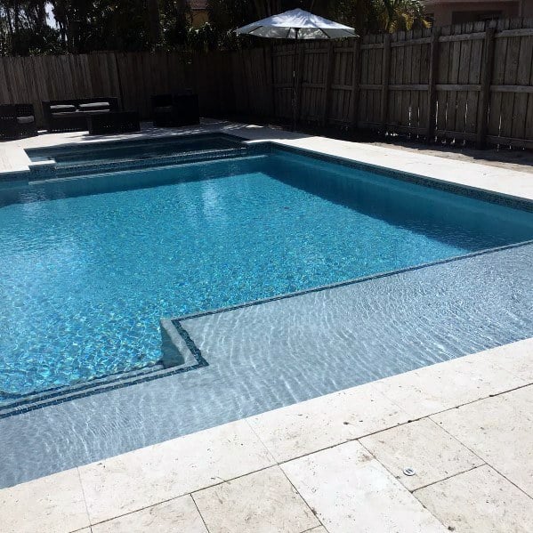 Rectangular pool with light blue mosaic tiles, bordered by a spacious stone deck and wooden fence.
