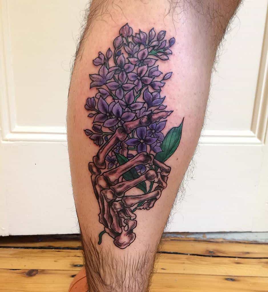 Bold tattoo of a skeleton hand gripping a bouquet of vibrant lilac flowers, blending gothic and botanical elements.