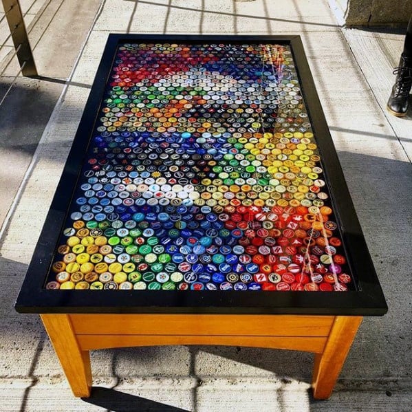 Table with multicolored bottle caps arranged under clear glass on a wooden frame, casting shadows on a concrete floor