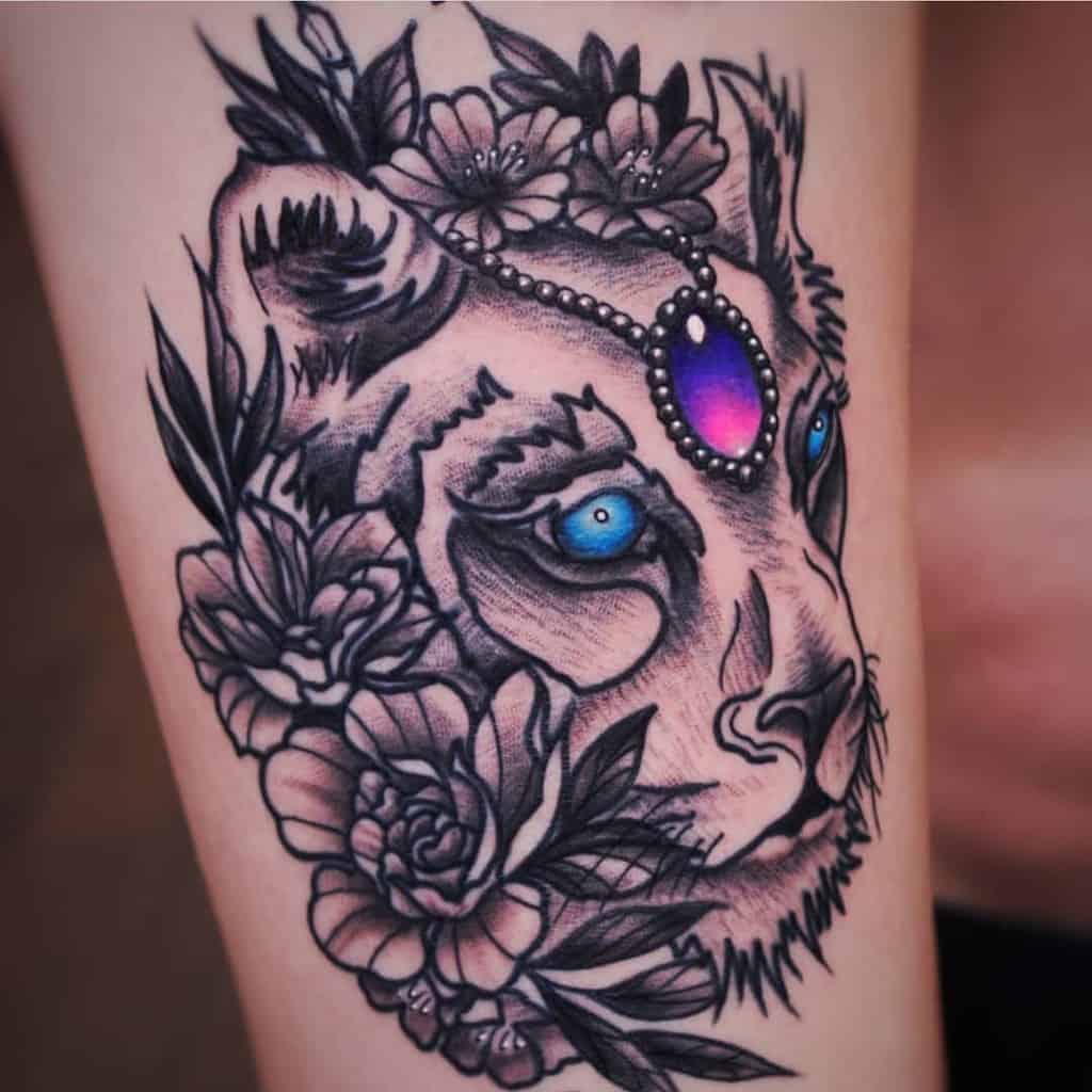Bold black and grey lioness tattoo with vibrant blue eyes, floral accents, and a jeweled headpiece featuring a glowing purple gemstone.