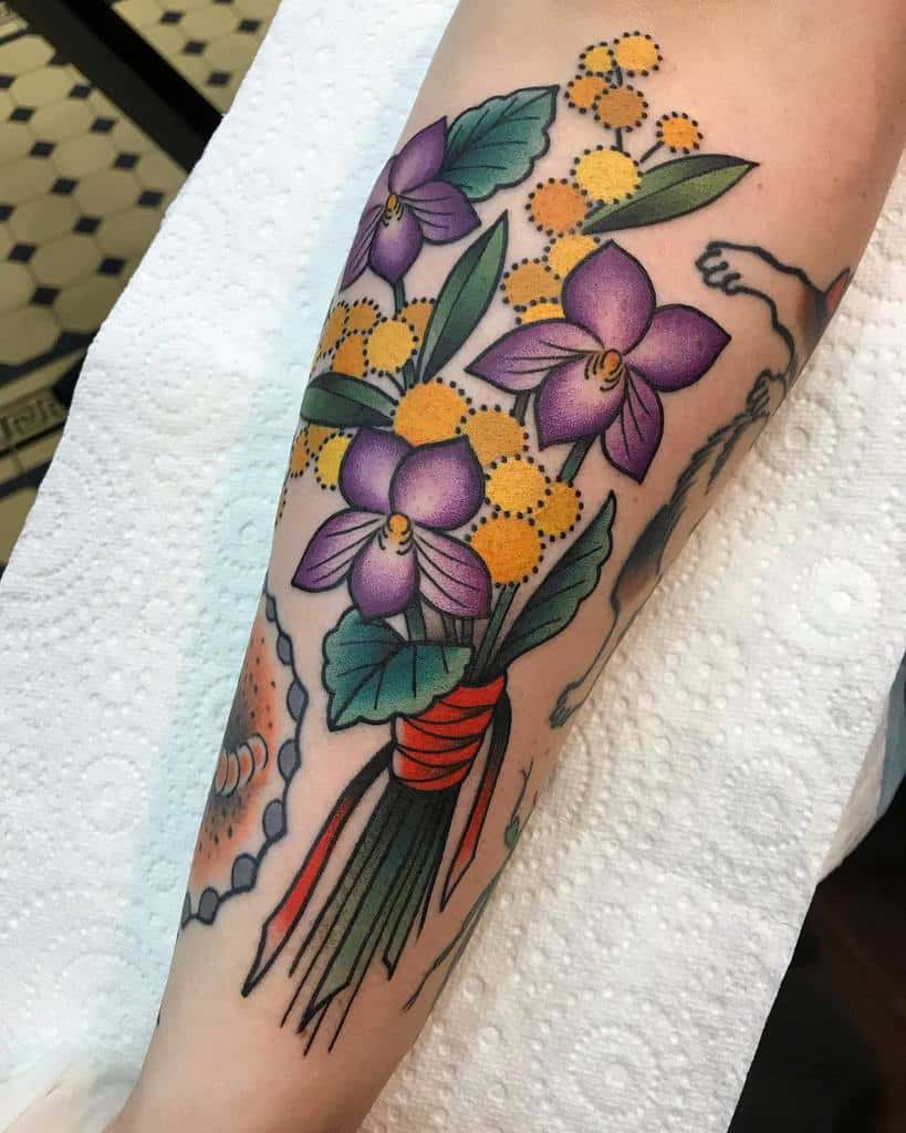 Bold and vibrant tattoo of a bouquet featuring purple violets, yellow mimosa flowers, and lush green leaves, tied together with a red ribbon in a traditional style