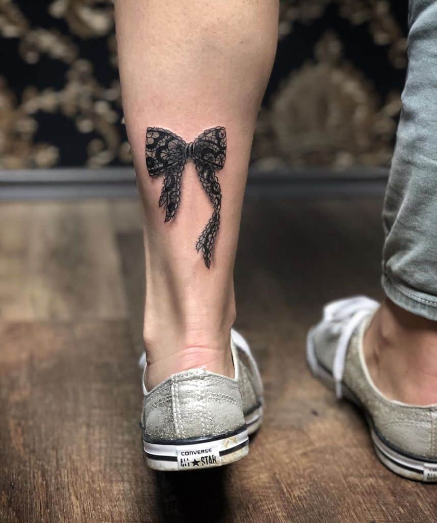 Delicate black lace bow tattoo on the back of the lower leg, featuring intricate shading and fine details for a realistic effect.