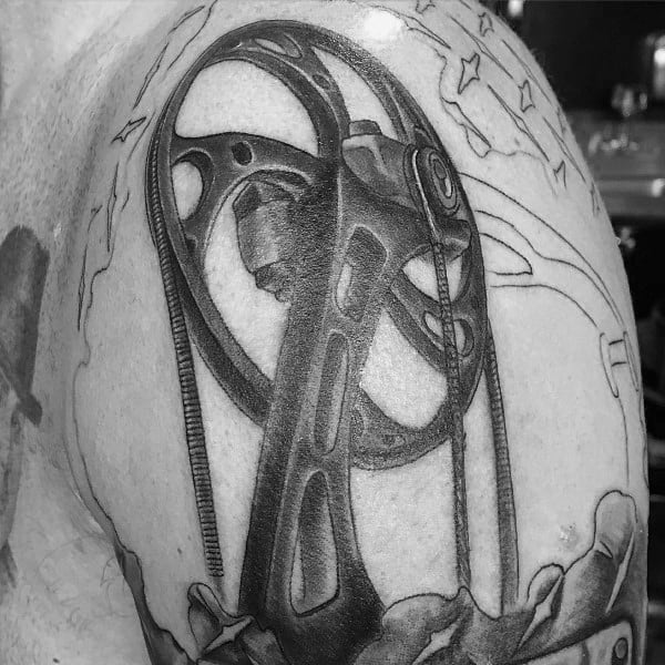 Detailed black and grey tattoo of a compound bow cam mechanism on the upper arm, surrounded by subtle background elements.