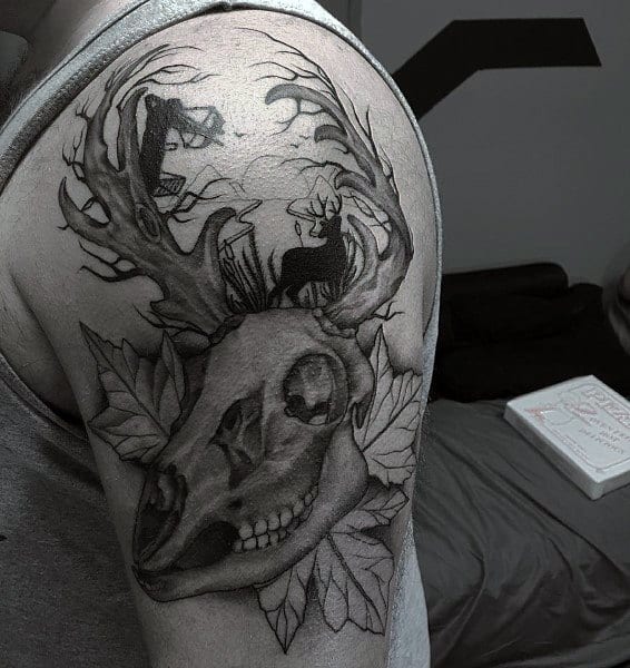 Black and grey shoulder tattoo of a deer skull with antlers forming a forest scene, including a bowhunter and a buck silhouette.
