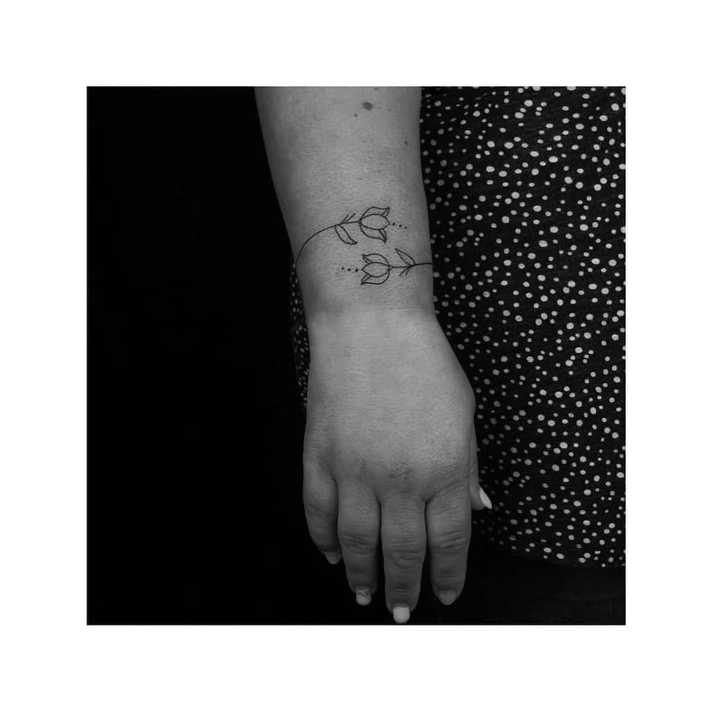 A minimal black-line tulip tattoo on the wrist, featuring two tulips connected by a thin, curved stem with small dot accents.