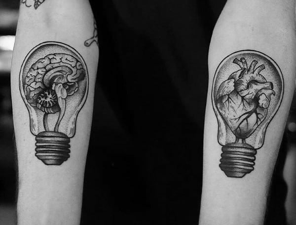 Matching forearm tattoos of a brain and a heart inside light bulbs, symbolizing the balance between logic and emotion.