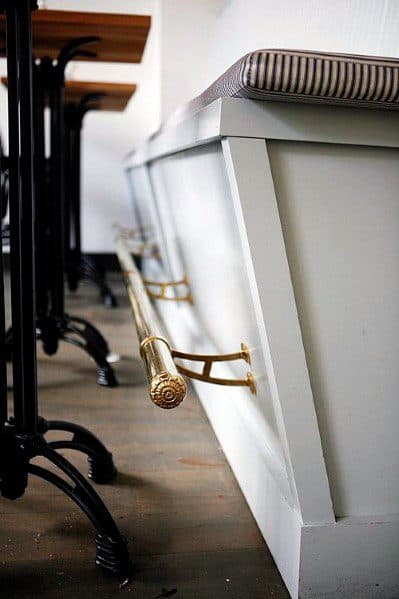 Elegant brass foot rail attached to a white bar with striped cushioned seating and wooden floors.
