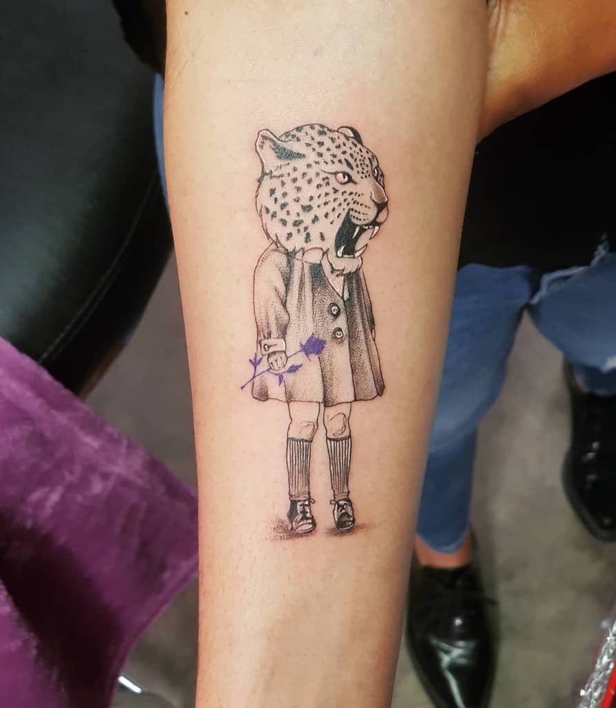 A unique tattoo of a jaguar's head on the body of a child in a coat, with a snarling expression and holding a flower, placed on the forearm for a quirky, artistic design.