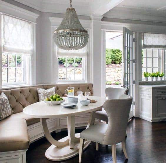 A cozy dining nook with a round table, tufted bench, chairs, and a beaded chandelier in a bright, airy room
