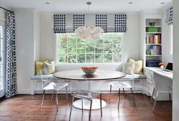 Bright dining nook with a round table, clear chairs, and a bowl of oranges next to a window seat and built-in shelves