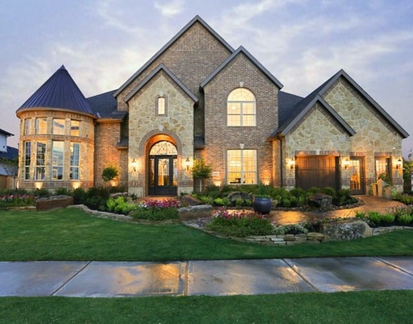 Stunning house with brick and stone exterior, turret-style bay windows, arched entry, and lush landscaping illuminated at dusk