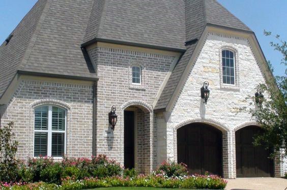 Charming home with light brick and stone exterior, steep roofline, arched garage doors, and vibrant flower-lined landscaping