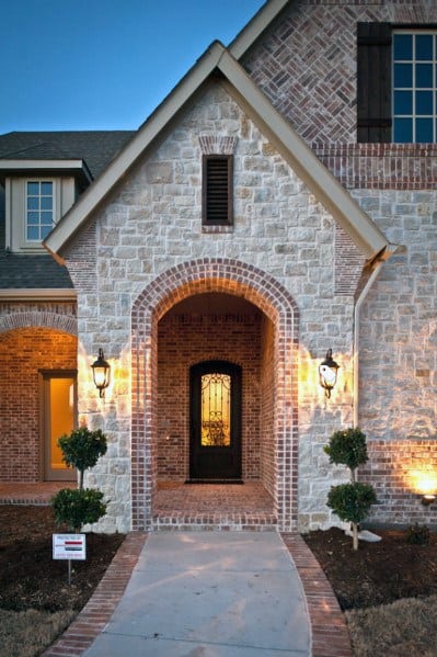Elegant entryway with arched brick and stone design, illuminated sconces, wrought iron door, and symmetrical topiary accents