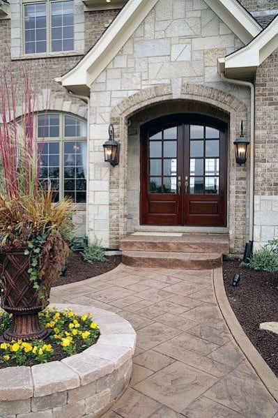 Inviting entryway with stone and brick cladding, double wooden doors, arched design, and a landscaped pathway with floral accents