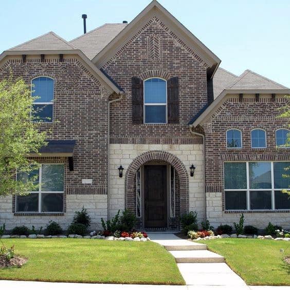 Classic two-story home with dark brick and light stone accents, arched entryway, rustic shutters, and a well-manicured front lawn
