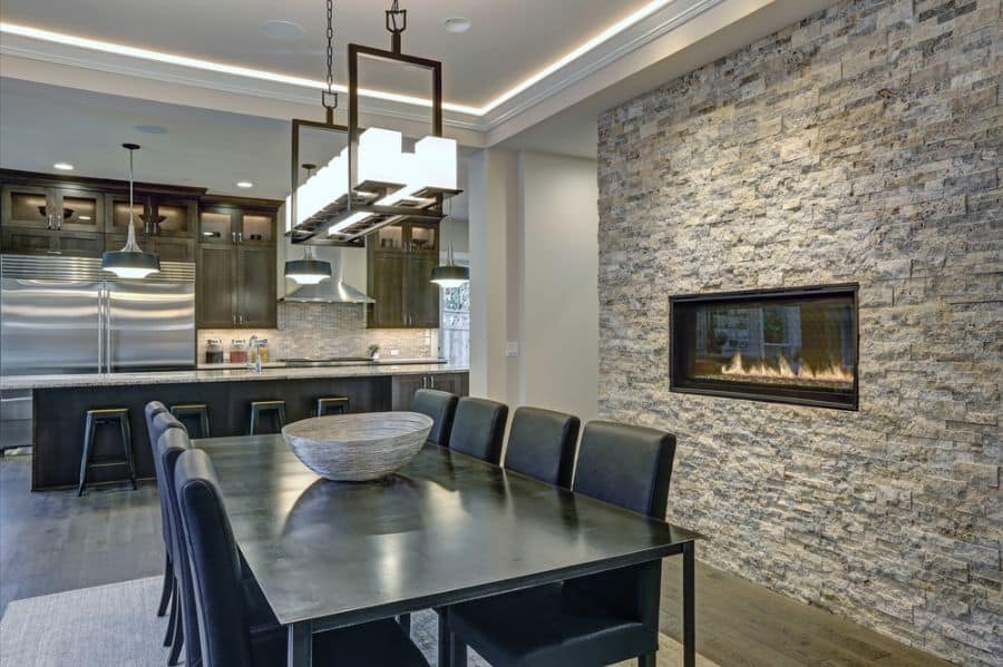 Modern dining room with an accent wall, long table, leather chairs, stone fireplace, and view of a sleek kitchen
