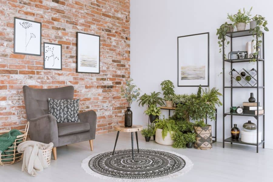 Cozy living room with a gray armchair, accent brick wall, plants, framed art, and a patterned round rug