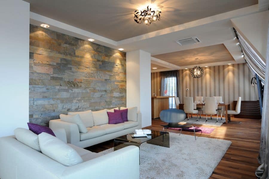 Modern living room with beige sofas, stone accent wall, and dining area in the background; warm lighting and sleek decor